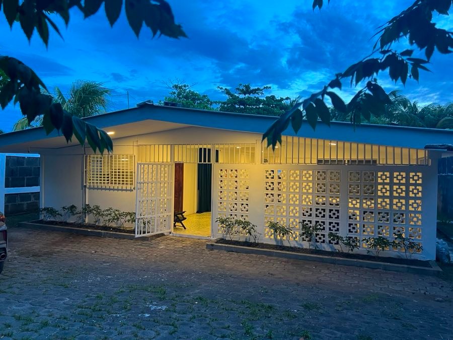 Fachada de Calm Beach House iluminada al anochecer, con celosías blancas y vegetación alrededor.