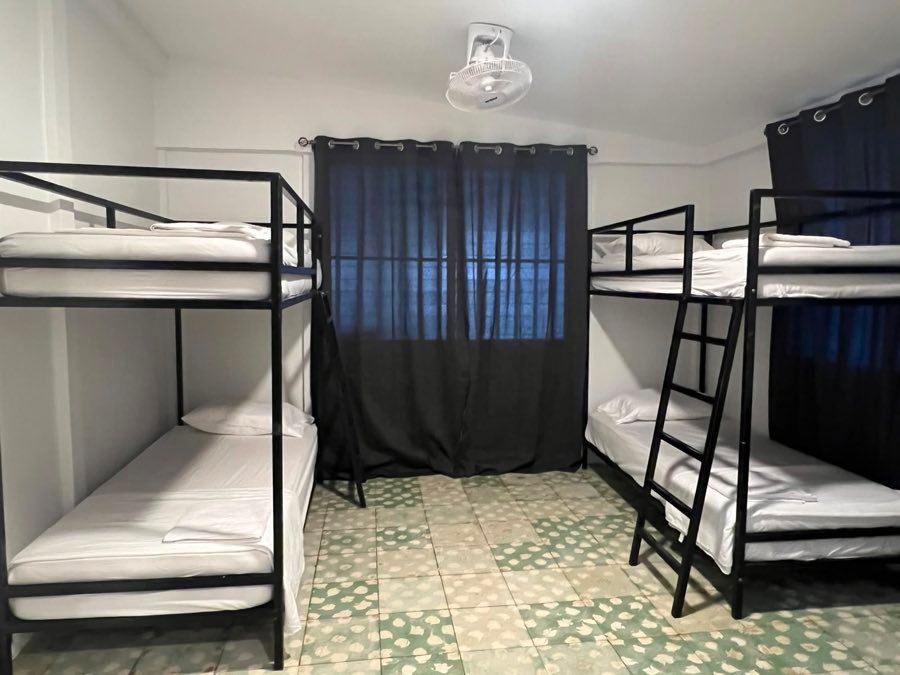 Habitación con literas de metal, ropa de cama blanca y cortinas oscuras.
